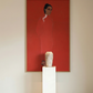 Artistic depiction of a figure against a red background with a white vase on a pedestal below. The Woman in Red – Portrait Canvas Wall Art | Large Luxury Print | Brume Art