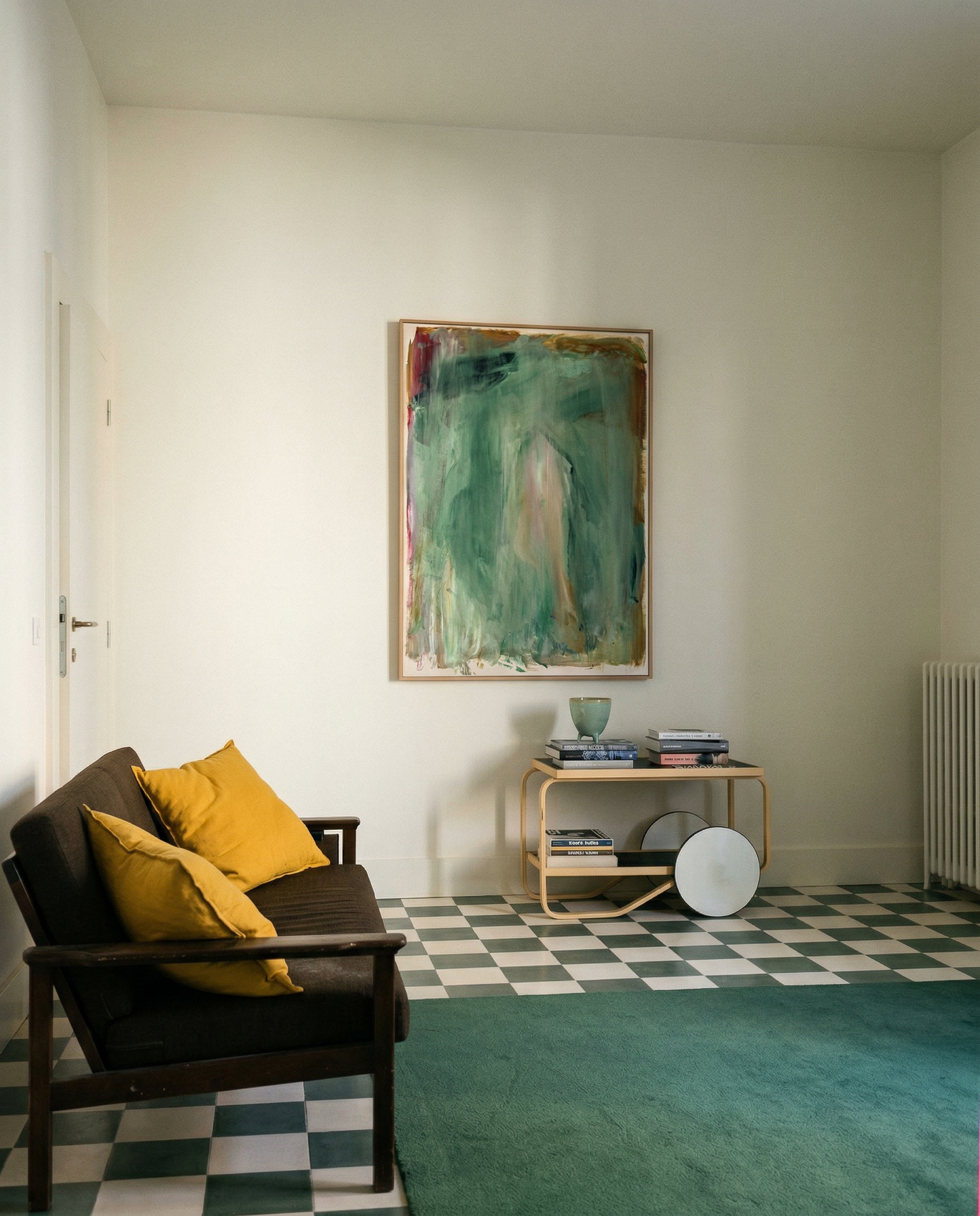 Living room with a dark sofa, yellow cushions, a checkered floor, and a large abstract painting on the wall. Abstract painting with green and brown tones on a white background Field green 2 – Abstract Canvas Wall Art | Large Luxury Print | Brume Art