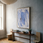 Nook with wooden bench, abstract blue painting, and potted plant against a light gray wall. Veil – Contemporary Abstract Canvas | Oversized Luxury Wall Art | Brume Art | Luxury Wall Decor | Large Canvas Wall Art | Oversized Wall Hanging-Brume art