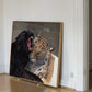 A framed painting of a black panther and a leopard in a warm-toned room, leaning against a white wall on a light wood floor.