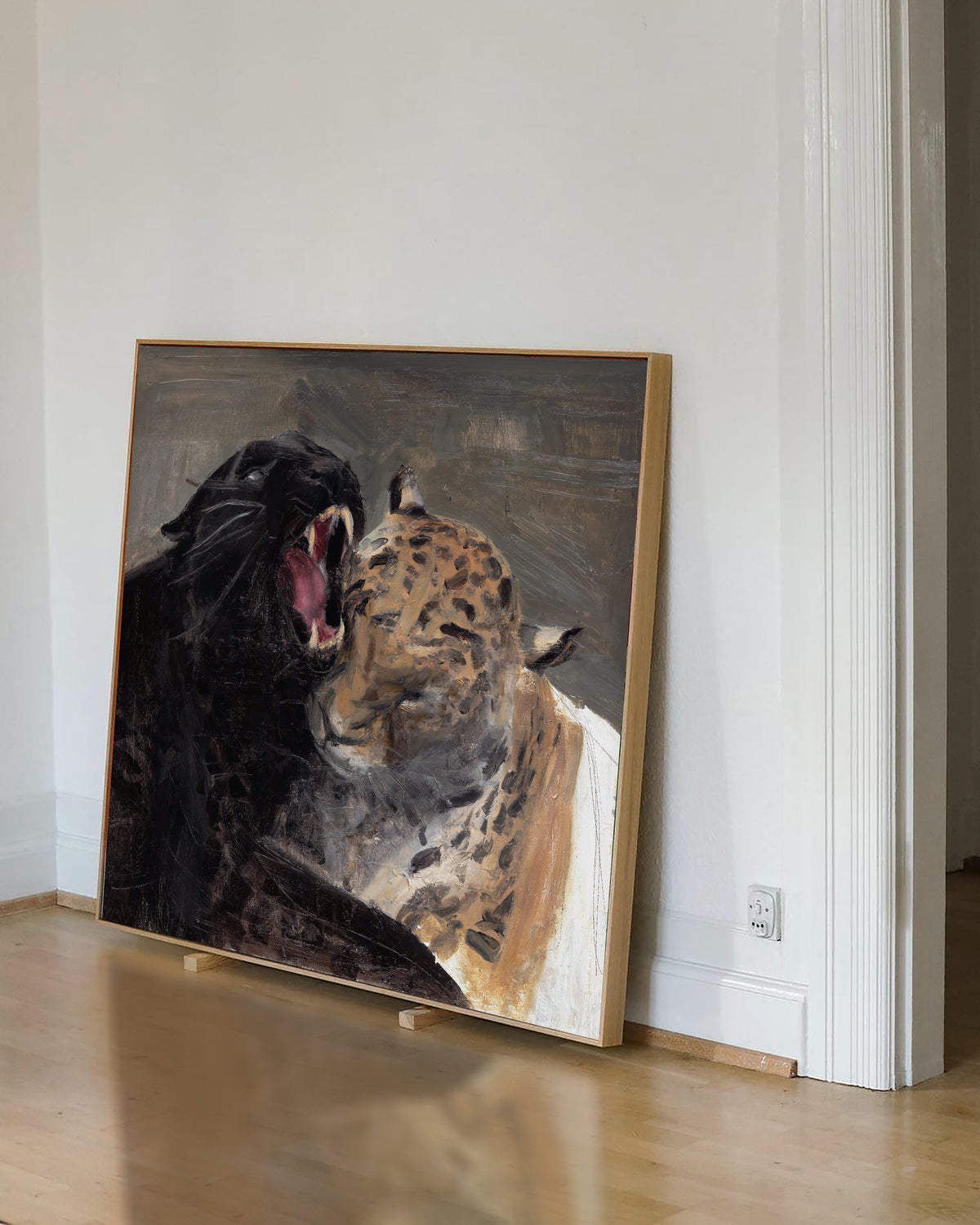 A framed painting of a black panther and a leopard in a warm-toned room, leaning against a white wall on a light wood floor.