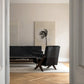 A modern interior scene viewed through a doorway, showing a black armchair and a black sofa in a minimalist living room with light wood flooring and a large abstract wall art piece.