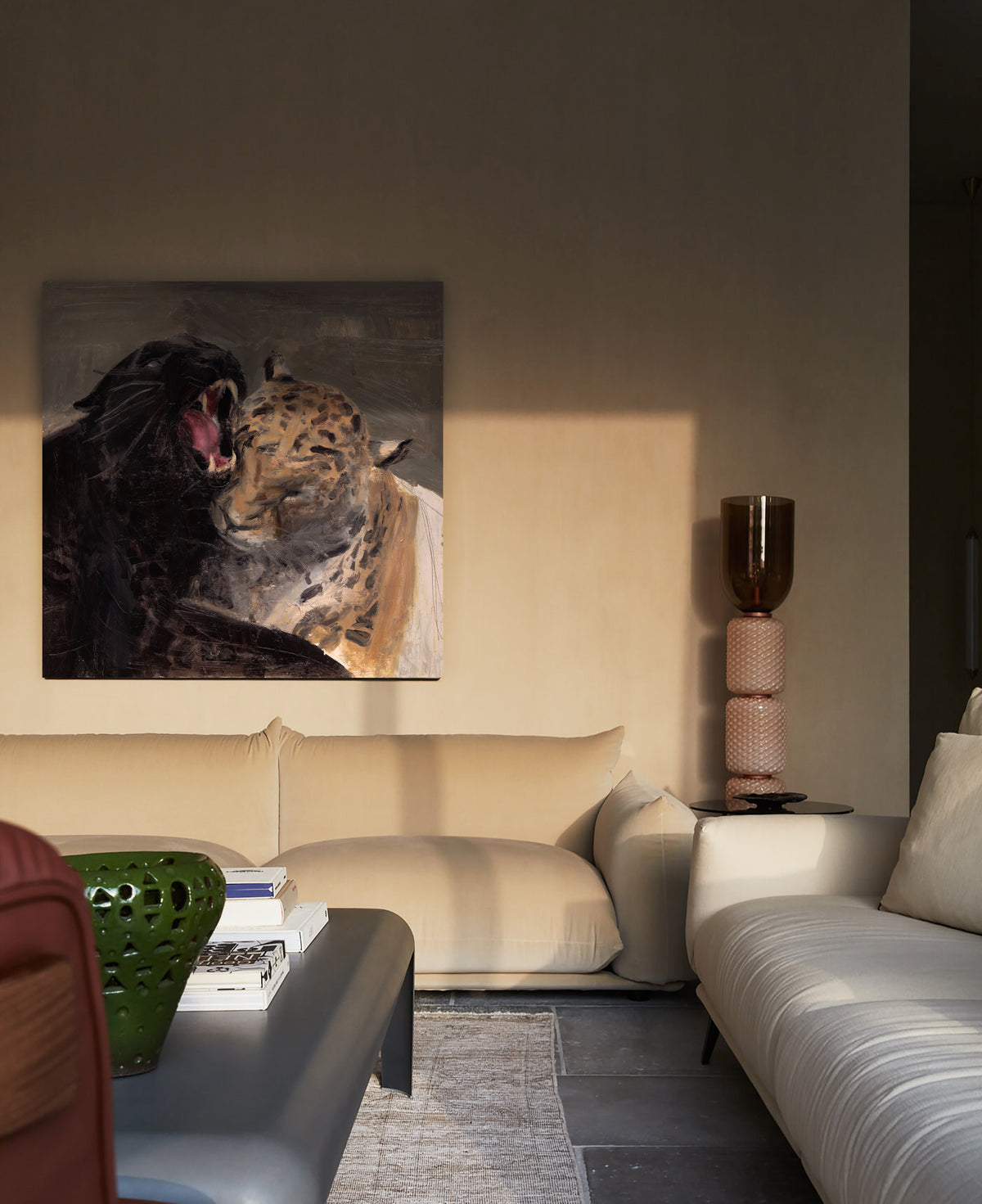 A square canvas painting of a leopard and a black cat hanging on a beige wall in a modern living room setting with a cream sofa and a gray coffee table.