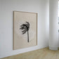 A framed abstract wall art print hangs on a white wall in a minimalist room with light wood flooring. The artwork features a black palm-like silhouette over a soft beige and off-white textured background, enclosed in a thin light wood frame.